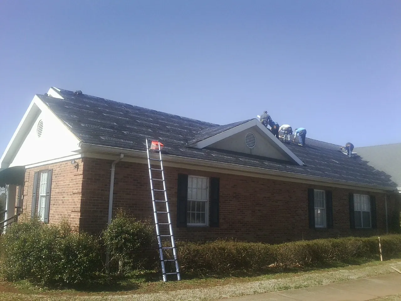 Crew tearing shingles off a brick ranch roof with a ladder set — Emergency Roof Repair work by our crew in Gueydan