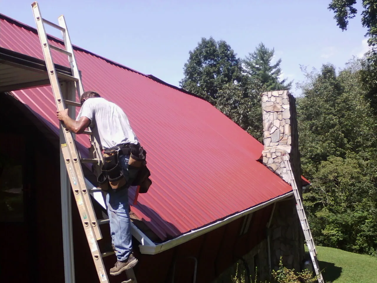 Roofer on a ladder beside a red metal roof and stone chimney — Soffit & Fascia Repair by Roofing Brothers in Gueydan
